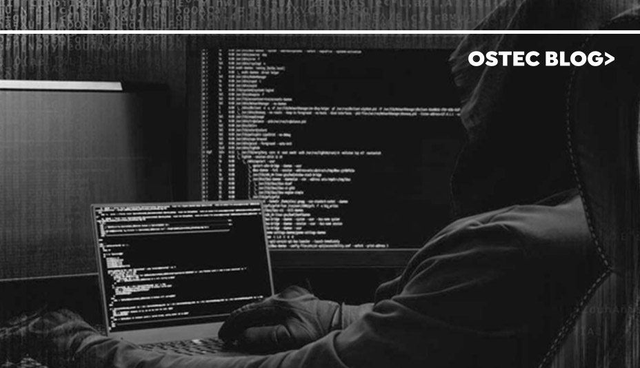 Homem sentado em uma sala escura com um notebook em sua frente e uma tela grande de computador ao seu lado, instalando um software pirata.