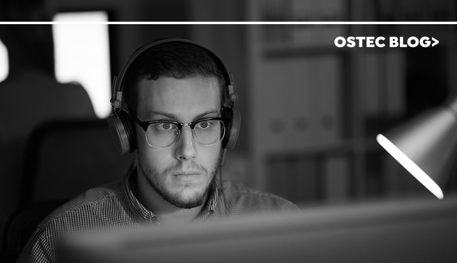 Homem de óculos e fone de ouvido sentado em frente a um computador trabalhando.